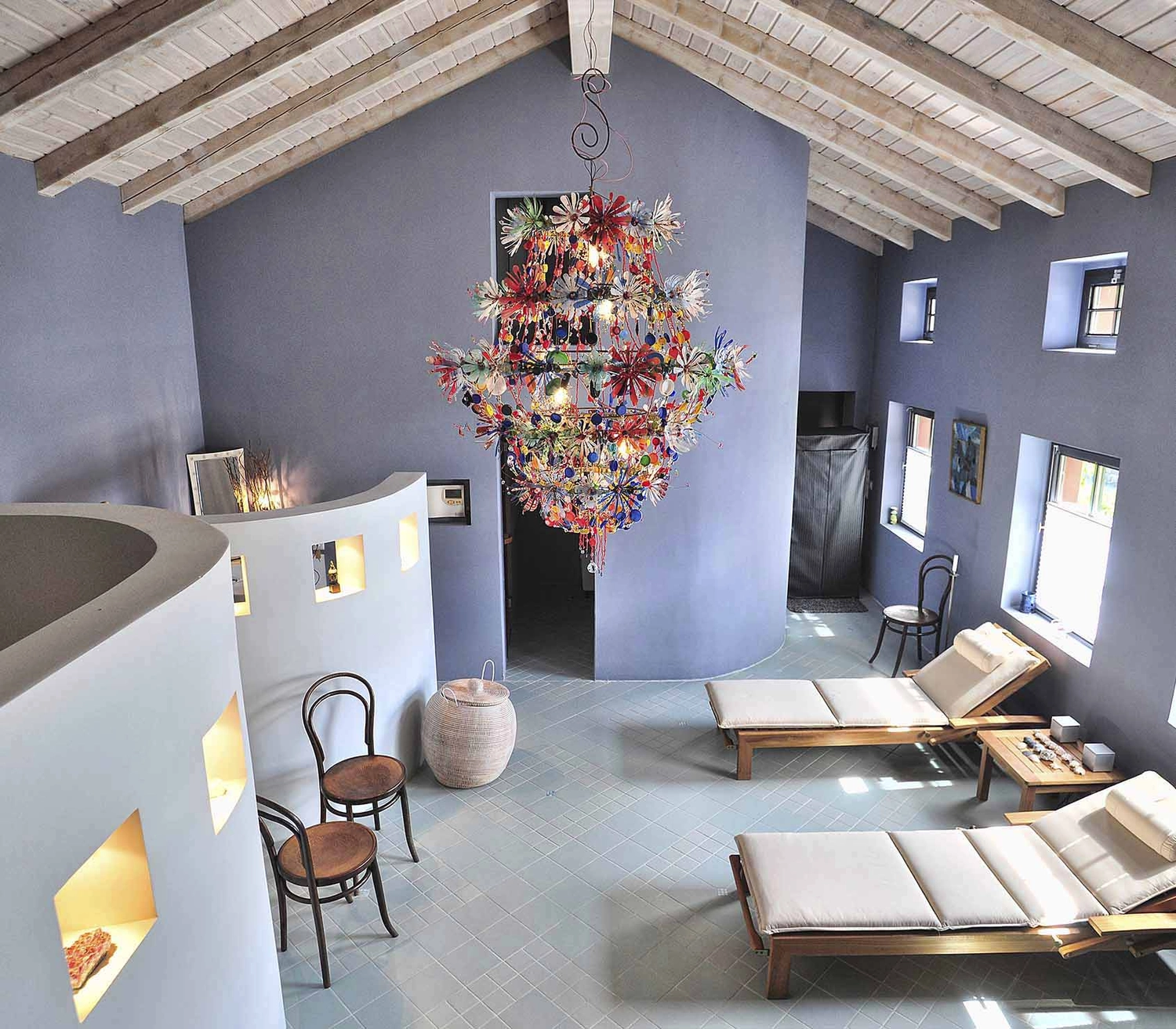 Atelier W - Kunst & Grafikdesign - Weitwinkelaufnahme eines geräumigen, modernen Zimmers mit hohen, gewölbten Decken und sichtbaren Holzbalken. Ein großer, mehrfarbiger Kronleuchter von Bleed, gefertigt aus filigranen Draht- und Glaselementen. Der Raum ist mit einem modernen Holzsessel mit weißen Kissen, einer runden, weißen Architekturnische und einem schlichten Beistelltisch aus Holz ausgestattet.